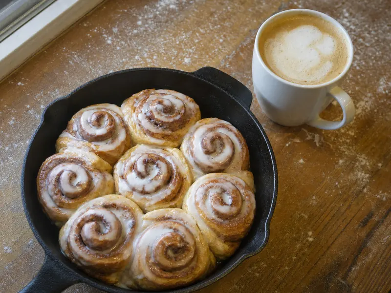 Glazed cinnamon rolls in a cast iron pan next to a cappuccino on a wooden counter.